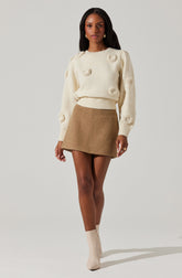 A woman is wearing a cream-colored, long-sleeved sweater with textured, circular details and a ribbed hem. She pairs it with a high-waisted, camel-colored, A-line mini skirt and matching cream-colored ankle boots.